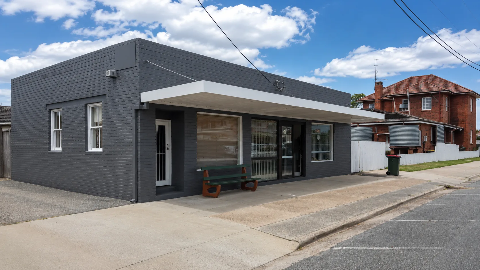 Grey brick commercial building with a freshly painted exterior, white trim, large street-facing windows and a bench outside under a blue sky.