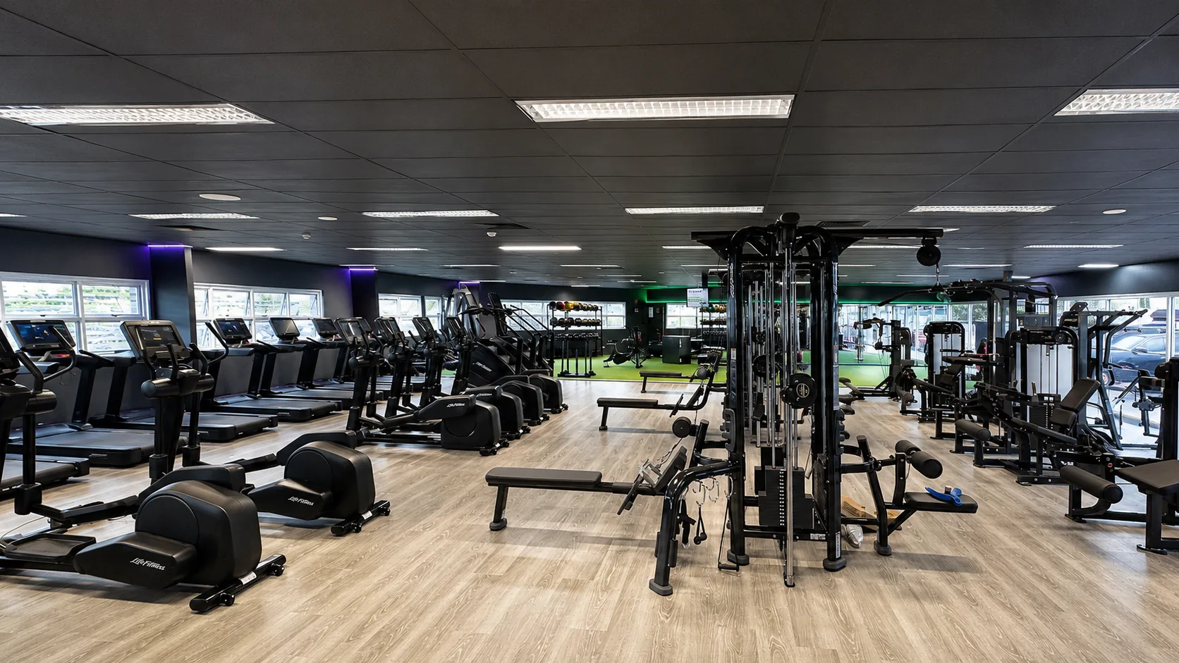 Spacious commercial gym with dark painted walls and ceiling, rows of cardio machines, cable machines, weight benches and green turf, showing interior painting work by Paint Gods.