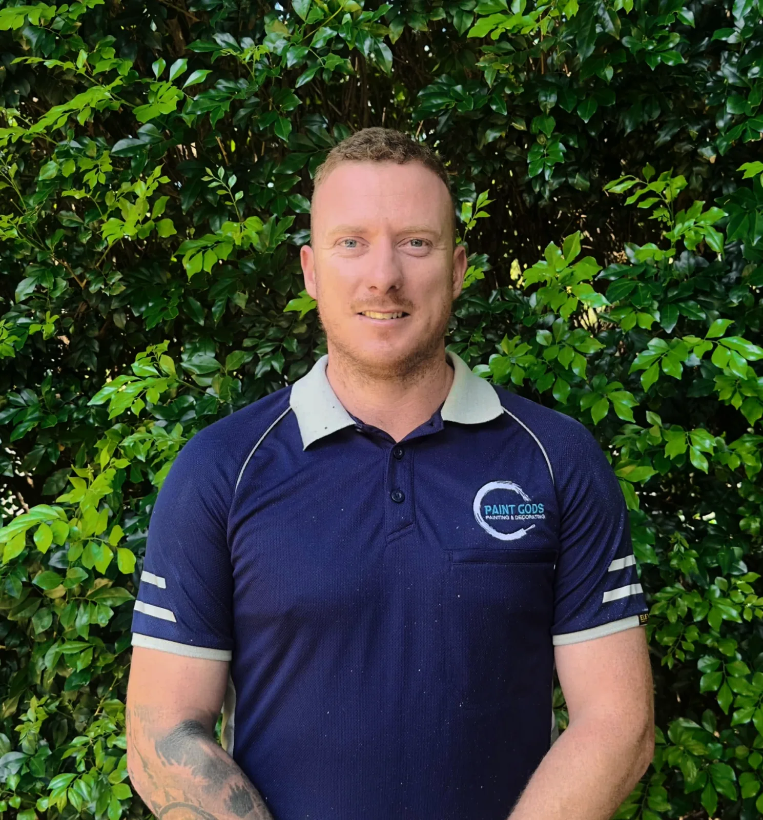 Ethan McMillan, Paint Gods owner and lead painter, standing in a navy branded polo in front of green foliage, with the shirt logo reading PAINT GODS PAINTING & DECORATING.